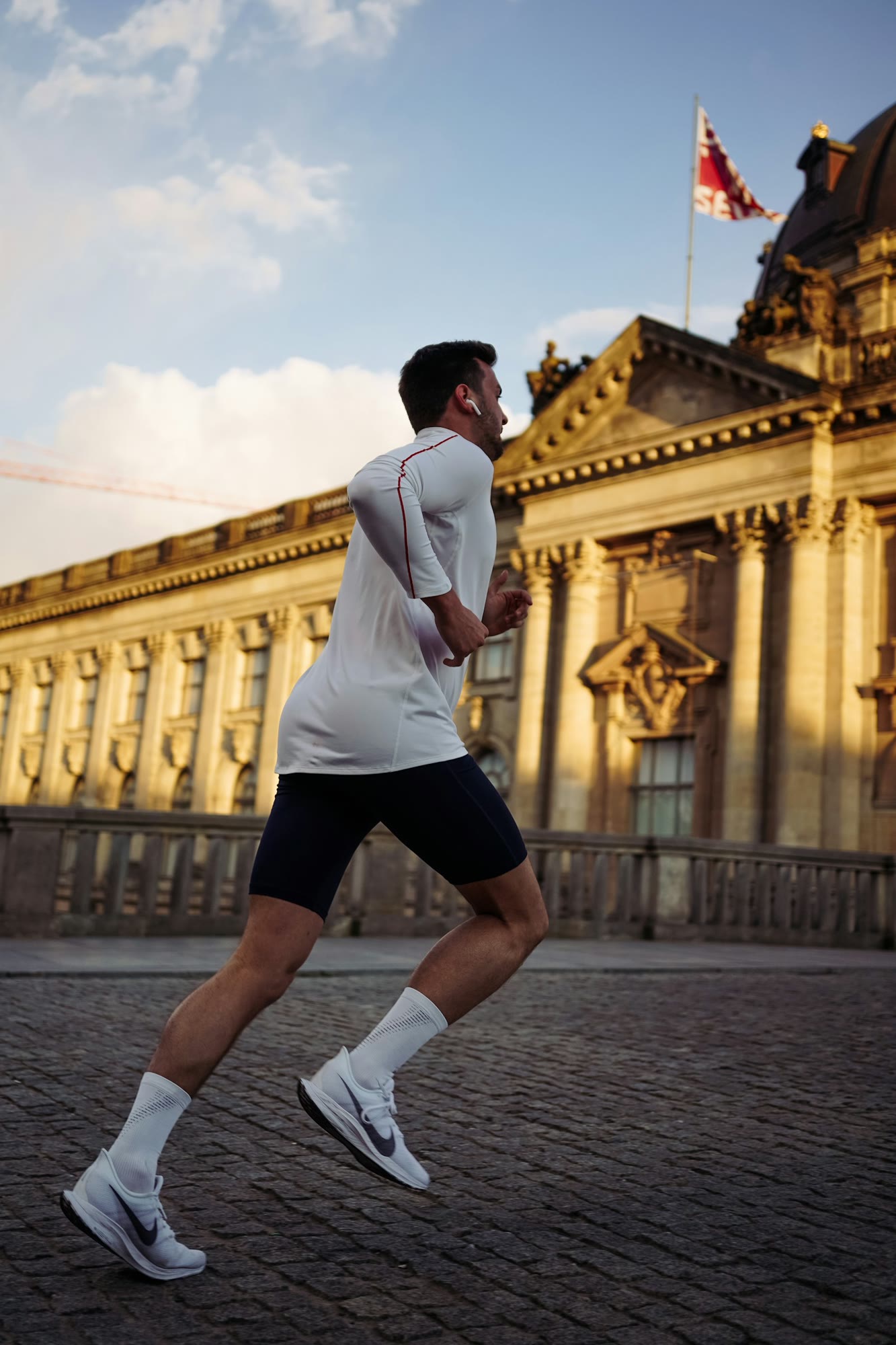 A person is running on a cobblestone street near an ornate, historic building with classical architecture. The runner is wearing athletic gear, headphones, and appears to be in motion under a clear sk