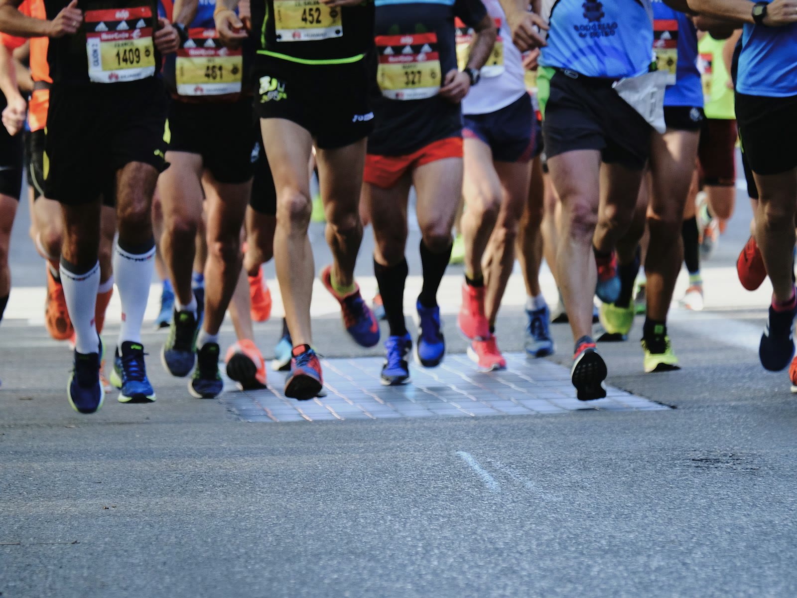 A group of runners is captured in motion during a marathon or race. The focus is on their legs and feet, showing a variety of running shoes and athletic wear. The ground is a paved road, and the runne
