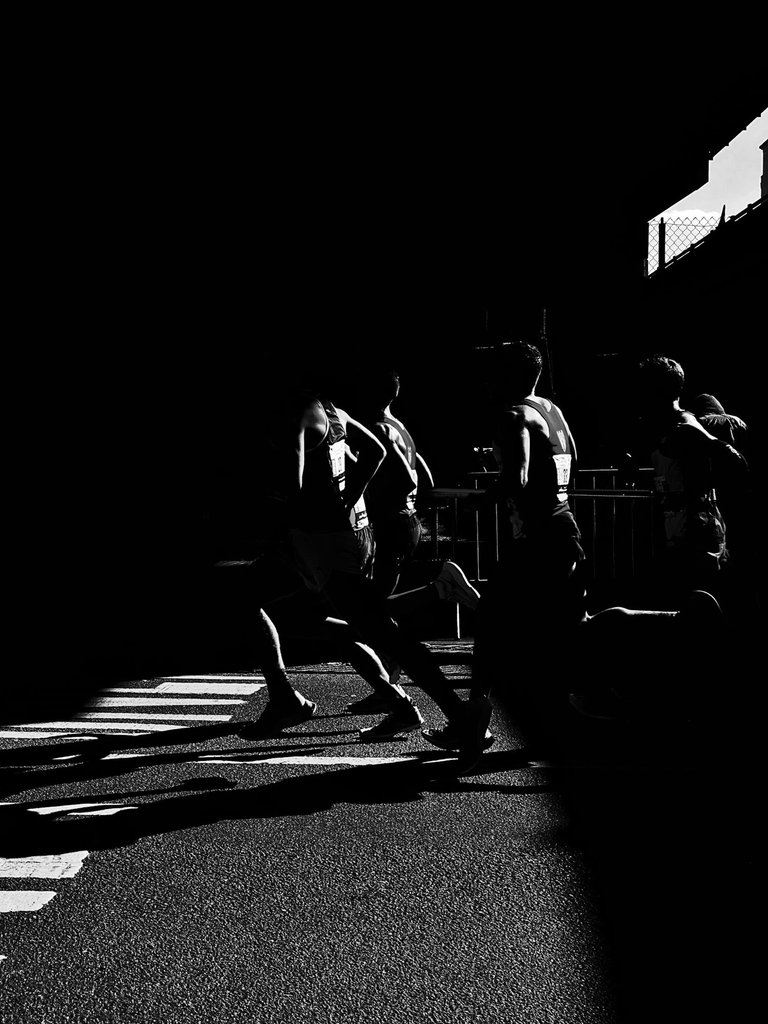 Silhouetted runners in a dramatic black and white scene, casting elongated shadows across a textured road surface. The high contrast highlights their dynamic movement and athletic form.