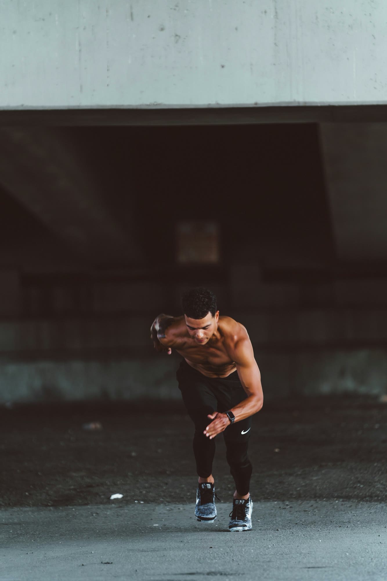 A muscular, shirtless man wearing black leggings and running shoes is captured in mid-stride, appearing to sprint forward. The setting is a dimly lit, urban environment with a concrete structure overh